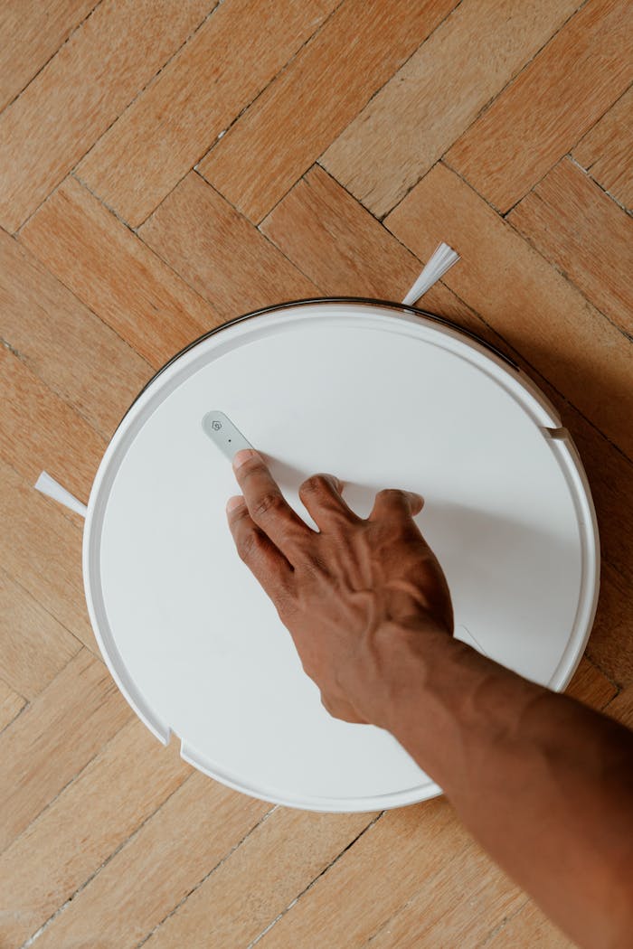 gallery-03 Close-up of a hand interacting with a robot vacuum cleaner on a parquet floor, showcasing modern cleaning technology.