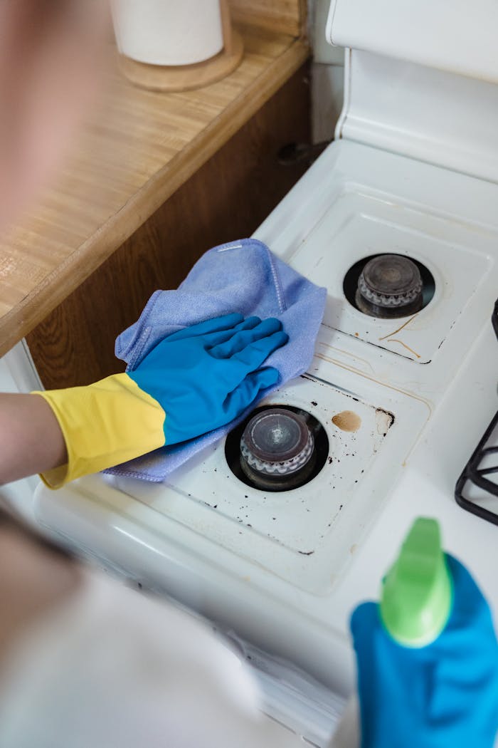 our-story A person wearing rubber gloves cleaning a dirty gas stove with a cloth and spray bottle.